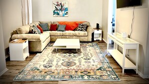 Una sala de estar con un sofá seccional color beige, una mesa de centro blanca, un tapete estampado, un ventilador de techo y un televisor de pantalla plana montado en la pared.