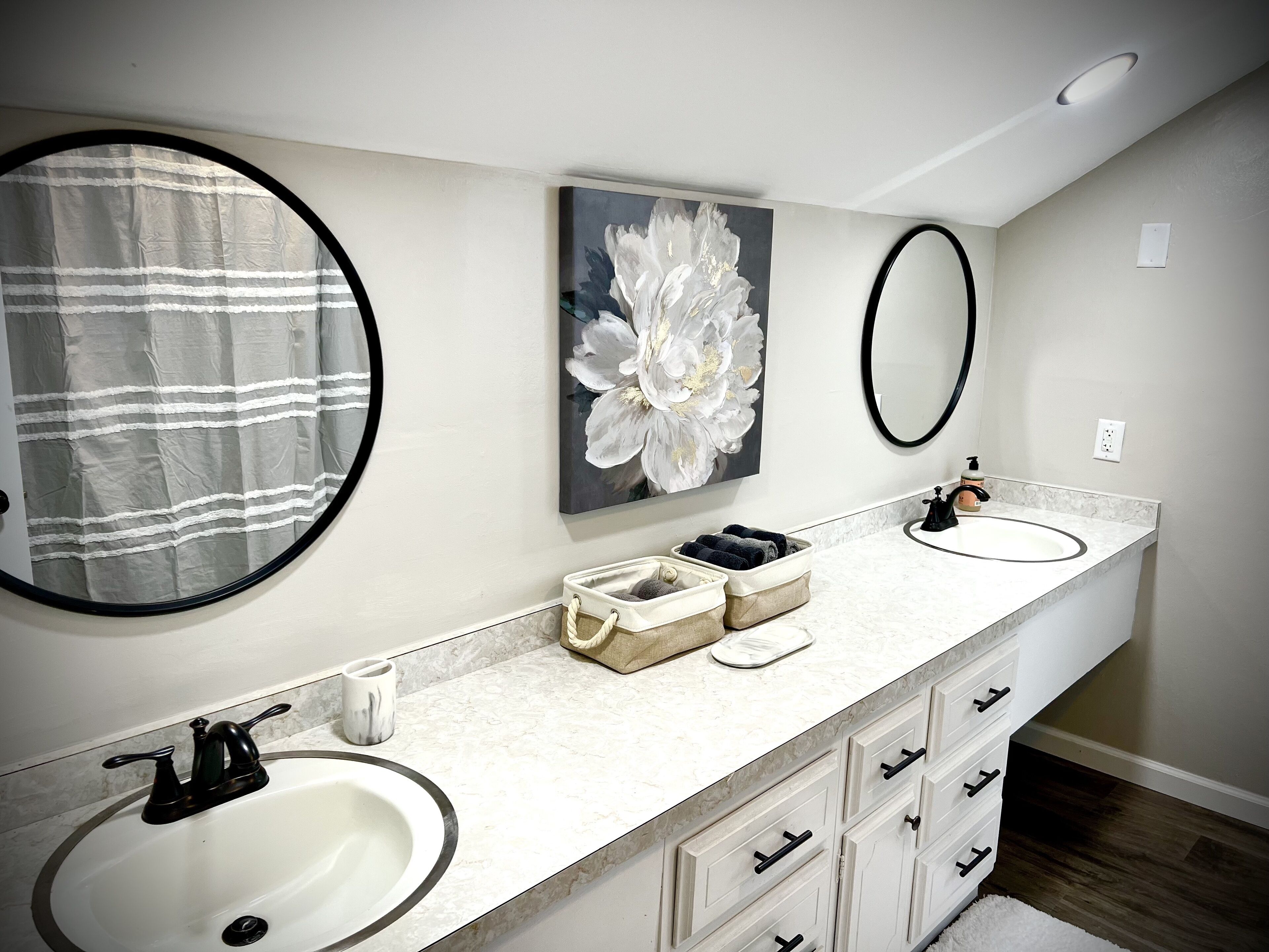 A modern bathroom with two sinks, a large mirror, a towel rack, and a floral artwork on the wall.