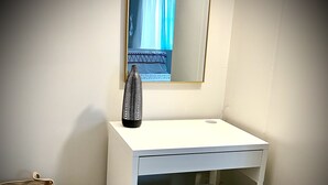 Una pequeña tocador blanco con un espejo arriba, un banco blanco, una cesta de mimbre y un cubo de basura blanco.