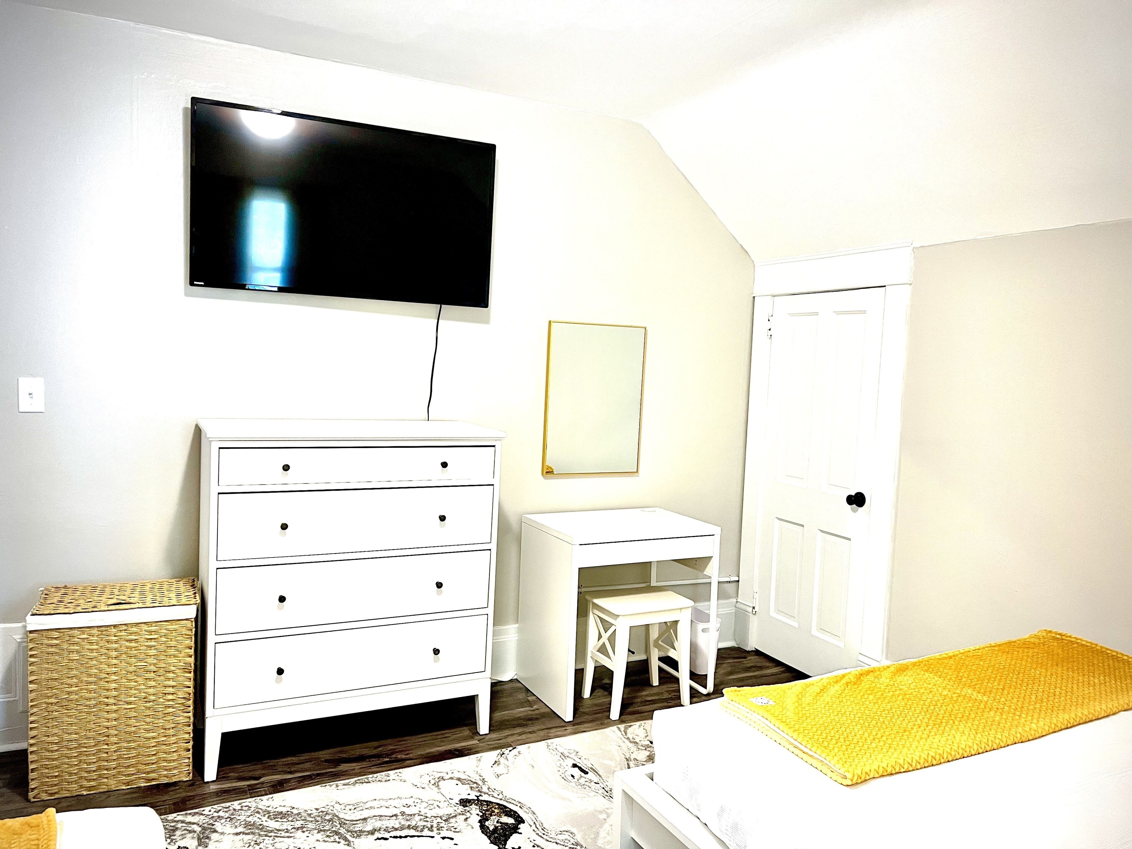 Ein Schlafzimmer mit Bett, Schminktisch, Schreibtisch, Stuhl, Fernseher und einer Tür.