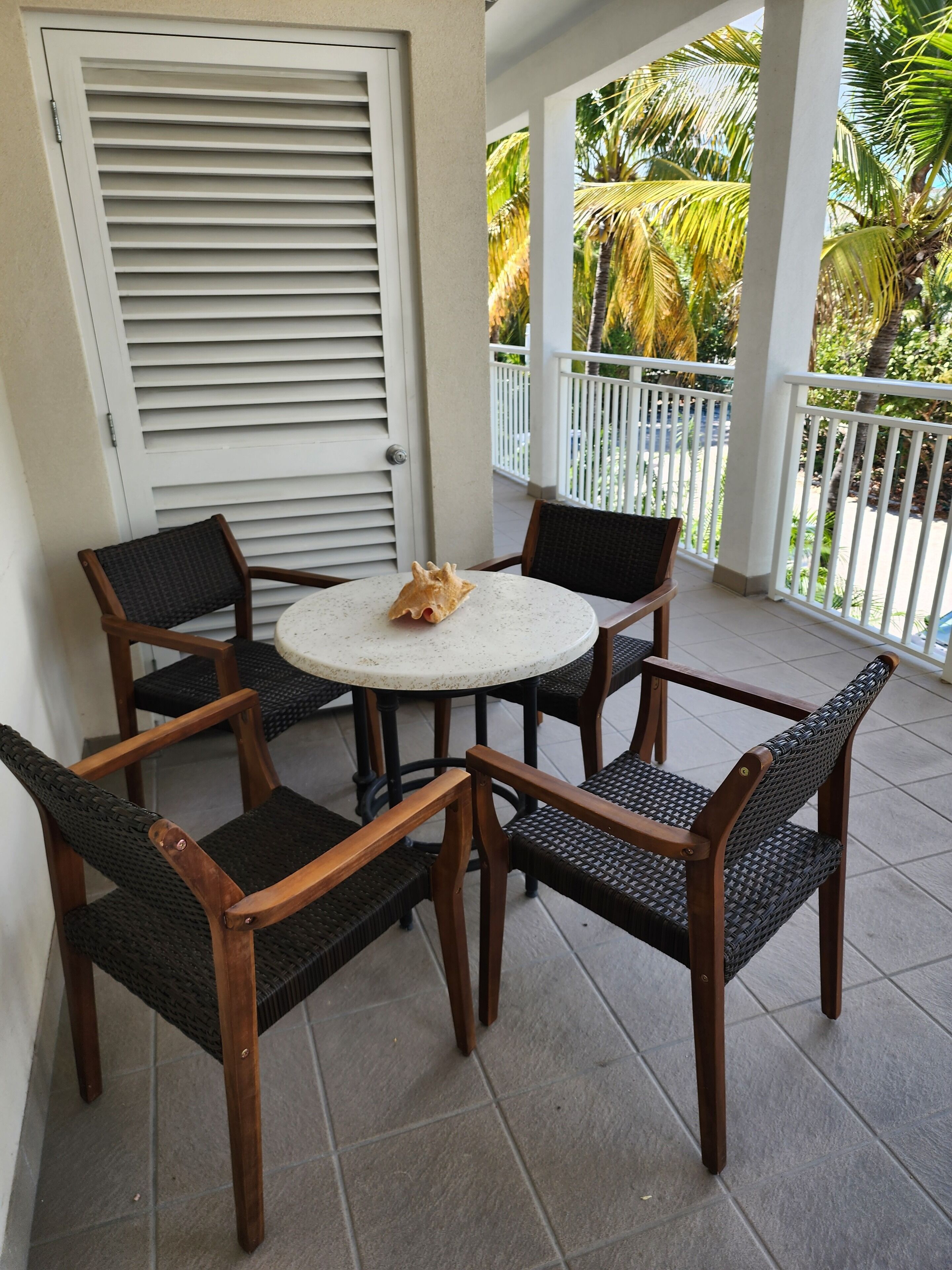 Un espace extérieur aménagé, abrité, avec une table ronde et quatre chaises, une porte à lattes horizontales et une vue sur des palmiers.