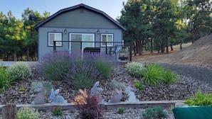 Una casa de fachada gris, un porche con un banco y un jardín con diversas plantas y elementos decorativos.