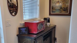 Una cocina con un microondas rojo, un reloj marcando las 7:00 de la noche, una ventana con persianas, un cuadro enmarcado y una planta en maceta.
