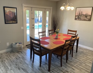 A dining area with a wooden table, red placemats, and six chairs. There is a door with glass panels, a chandelier, and two framed pictures on the walls.