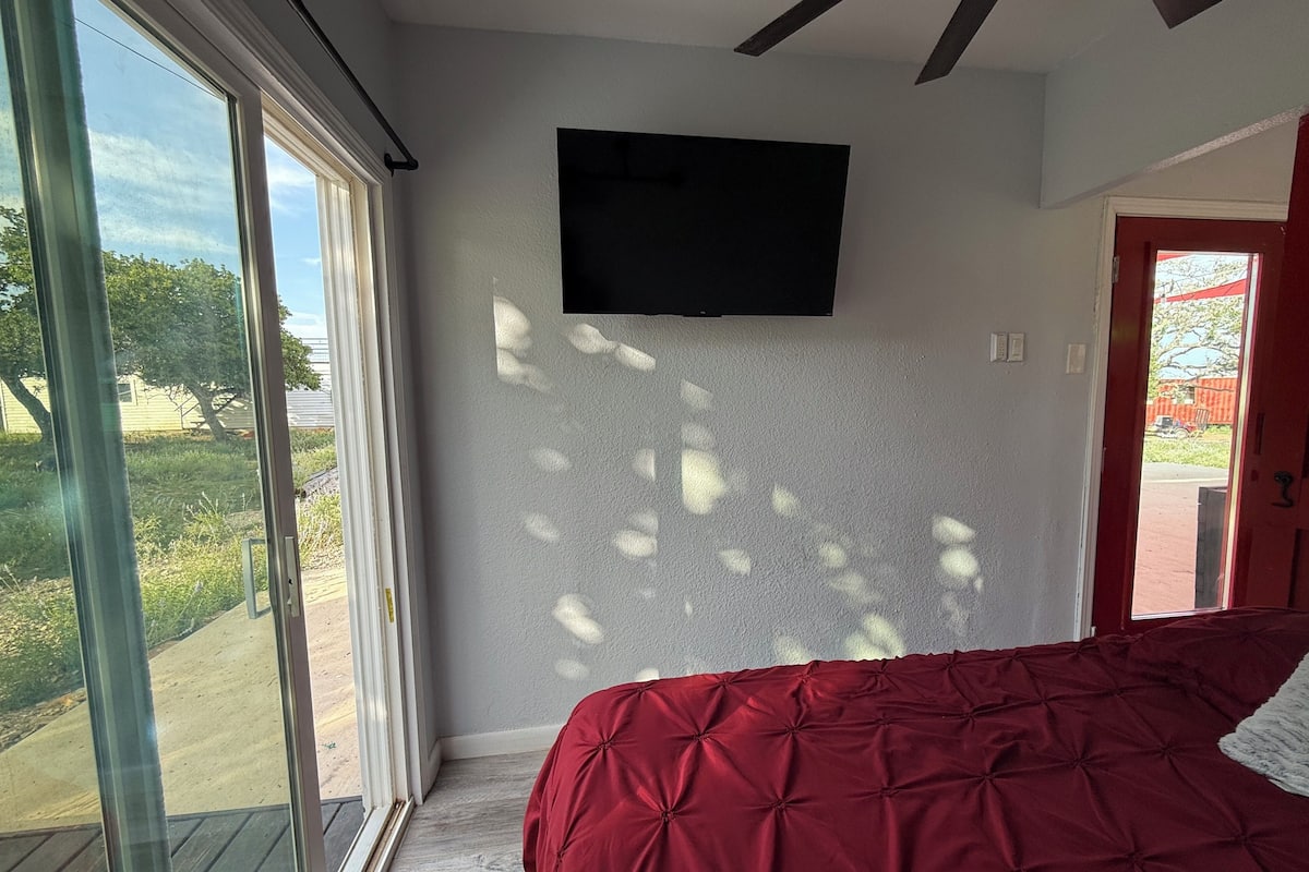 Un dormitorio con un ventanal grande, una cama con cubierta roja y un televisor de pantalla plana montado en la pared.