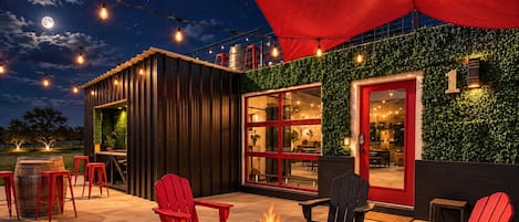 An outdoor seating area with a fire pit, red chairs, and string lights against a backdrop of a building with a green wall and red accents.
