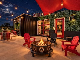 Een terras met buitenmeubels, een vuurkuil, rode stoelen en lichtslingers, met op de achtergrond een gebouw met een groene wand en rode accenten.