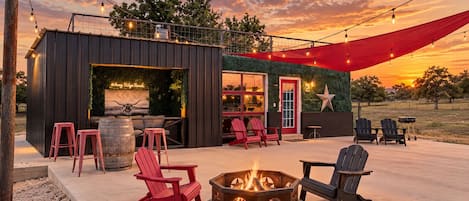A rustic outdoor seating area with Adirondack chairs, a fire pit, and string lights against a sunset backdrop.