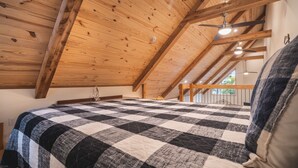 Korkeakattoinen huone, jossa on näkyvillä puiset kattopalkit, ruutukuviollinen peitto sängyllä ja kattotuuletin.