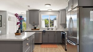 A modern kitchen with grey cabinets, a stainless steel refrigerator, and a window with a view of greenery. - Ridge View Retreat with Mtn Views & Hot Tub (Port Angeles)
