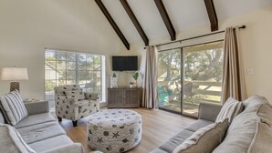 Ein Wohnzimmer mit hoher Decke, einer Couch, einem runden Hocker, einem Fernseher und einer Glastür zum Außenbereich.