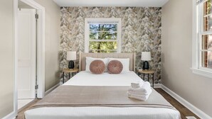 A bedroom with a large bed, two bedside tables with lamps, a window with a view of trees, and a door.