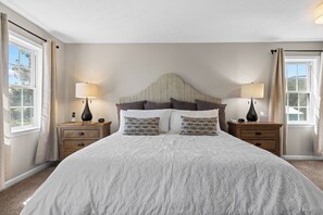 A bedroom with a large bed, two bedside tables, a headboard, and two windows with curtains.