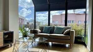 Un salon moderne avec un canapé, une table basse en verre et une vue sur les immeubles à travers de grandes fenêtres.