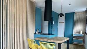 Une cuisine moderne avec une table en bois et des chaises jaunes, un luminaire suspendu noir et un mur d’accent bleu.