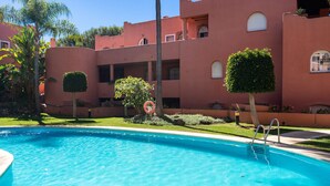 Una piscina con agua cristalina azul, rodeada por un edificio de fachada roja, palmeras y un césped bien cuidado.