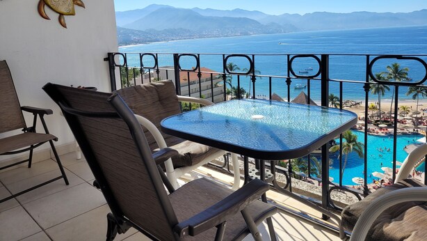 Un balcón con una mesa de vidrio y dos sillas, con vista a una playa y una zona de piscina.