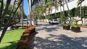 Un sendero pavimentado bordeado de palmeras y flores rojas, que conduce a un coche blanco estacionado cerca de un edificio con tejado de tejas rojas.