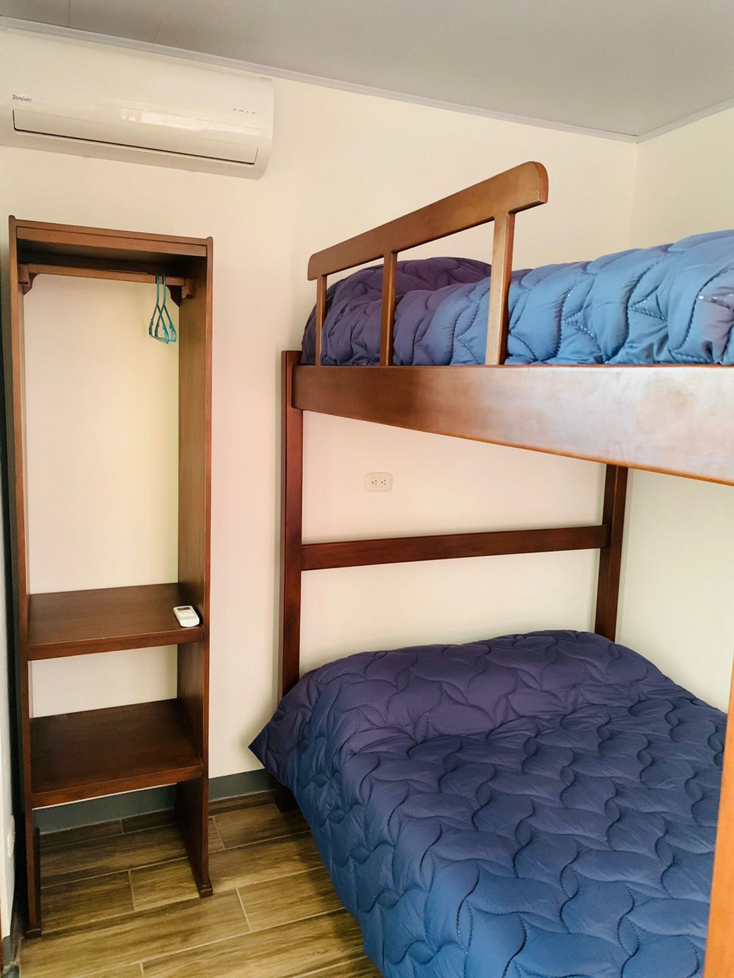 A bunk bed with a wooden headboard and a lower bed with a blue quilt. There is a wooden shelf unit to the left and a white air conditioning unit above.