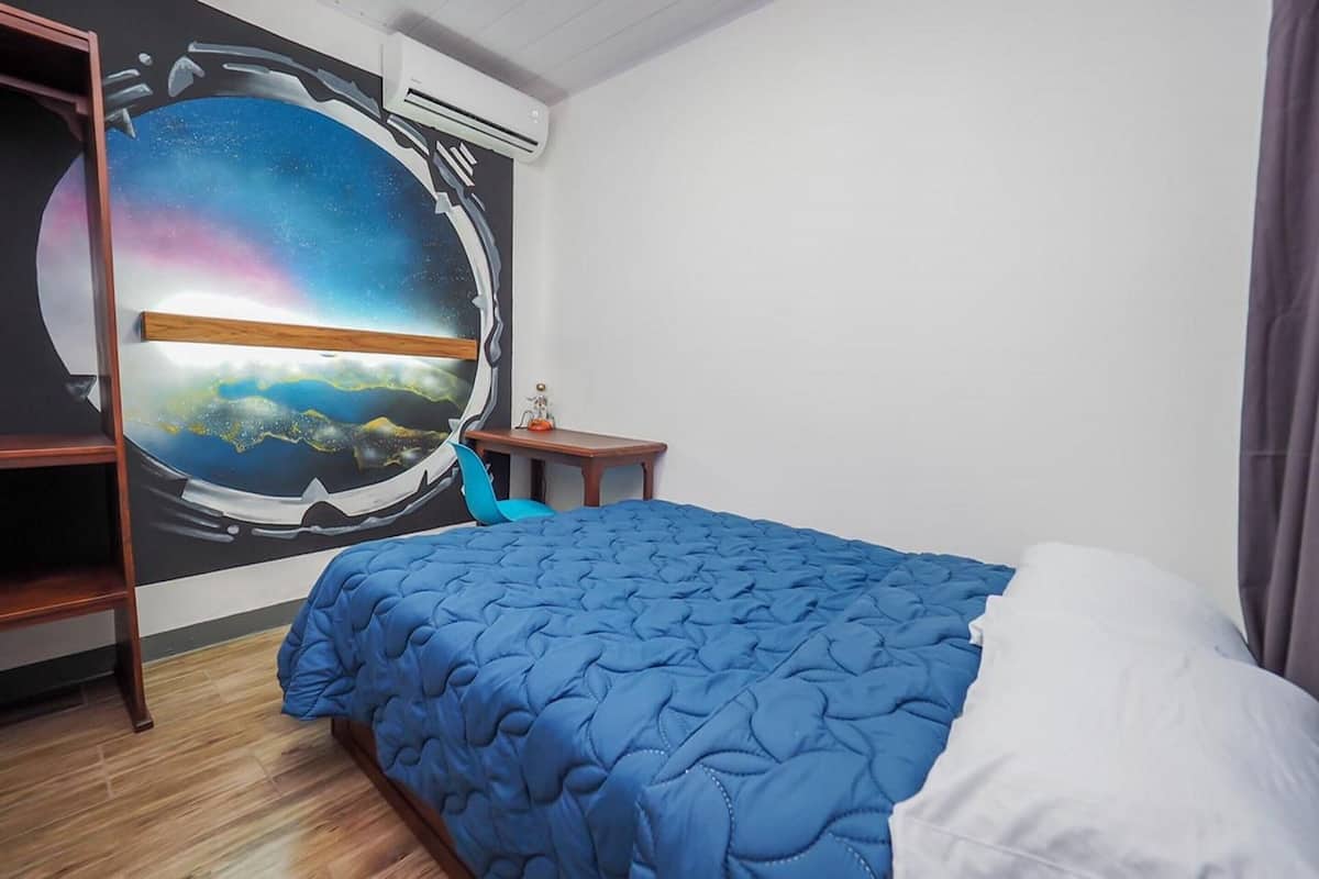Habitación de hotel con un mural grande de una escena espacial en la pared, una cama con colcha azul, una mesita de noche de madera y una pequeña mesa con un objeto decorativo.