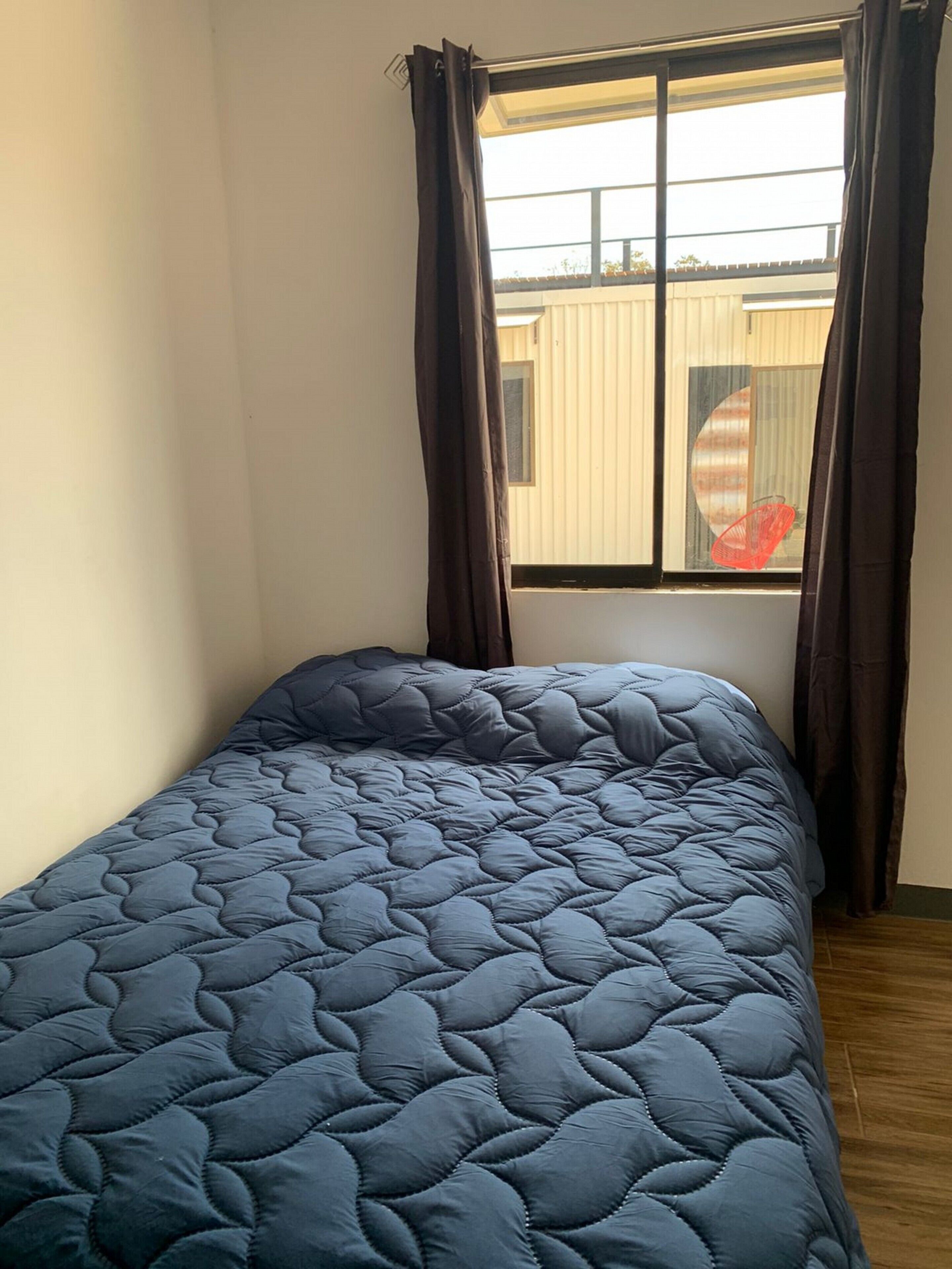 A single bed with a blue quilt in a room with a window and curtains.