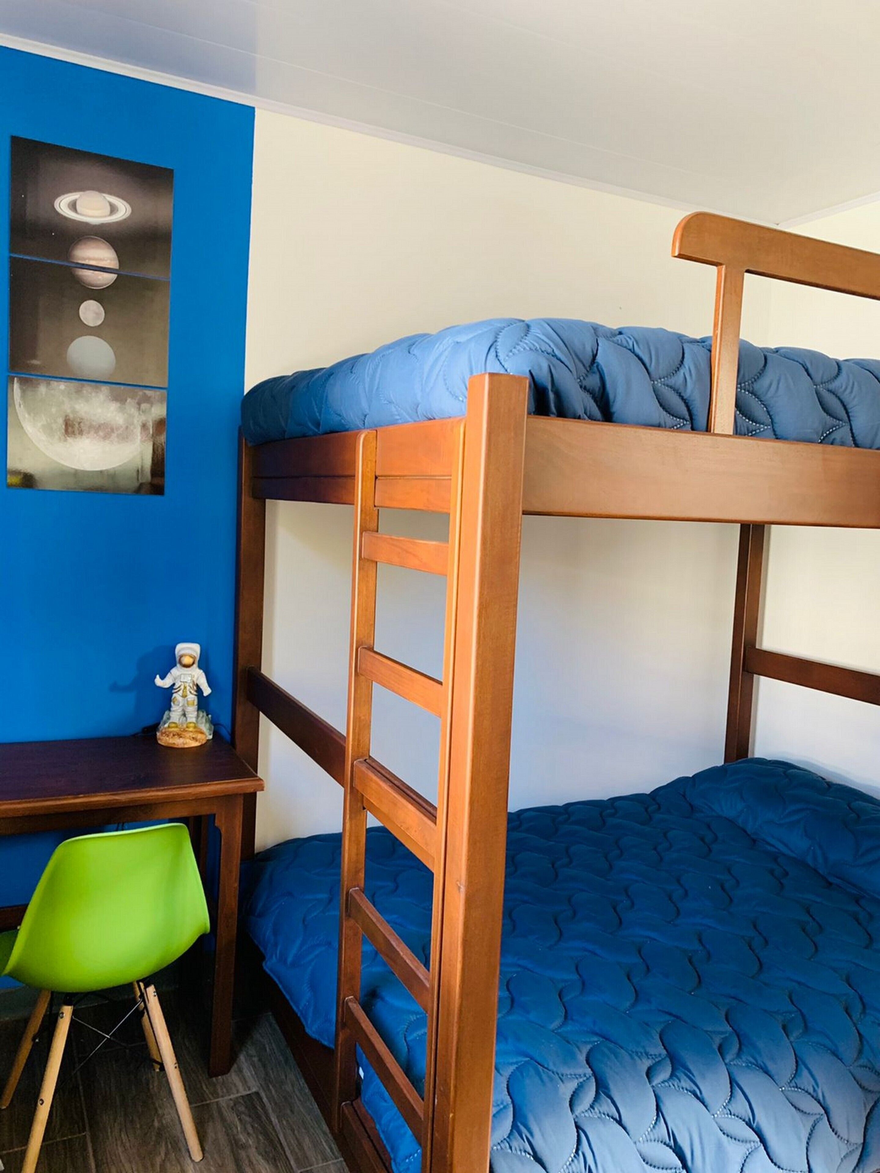 Un lit superposé avec une structure en bois et une literie bleue, un bureau avec une chaise, et un mur orné d’un tableau encadré.