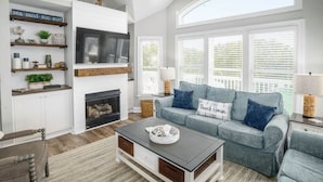 Ein Wohnzimmer mit Kamin, einem grauen Sofa mit 'Beach House'-Kissen, einem Couchtisch und einem Bücherregal mit Dekoration.