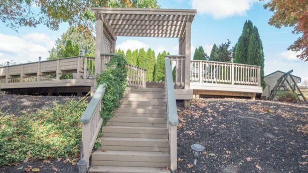 Una escalera de madera que lleva a una pérgola con barandilla, rodeada de árboles y un cielo despejado.