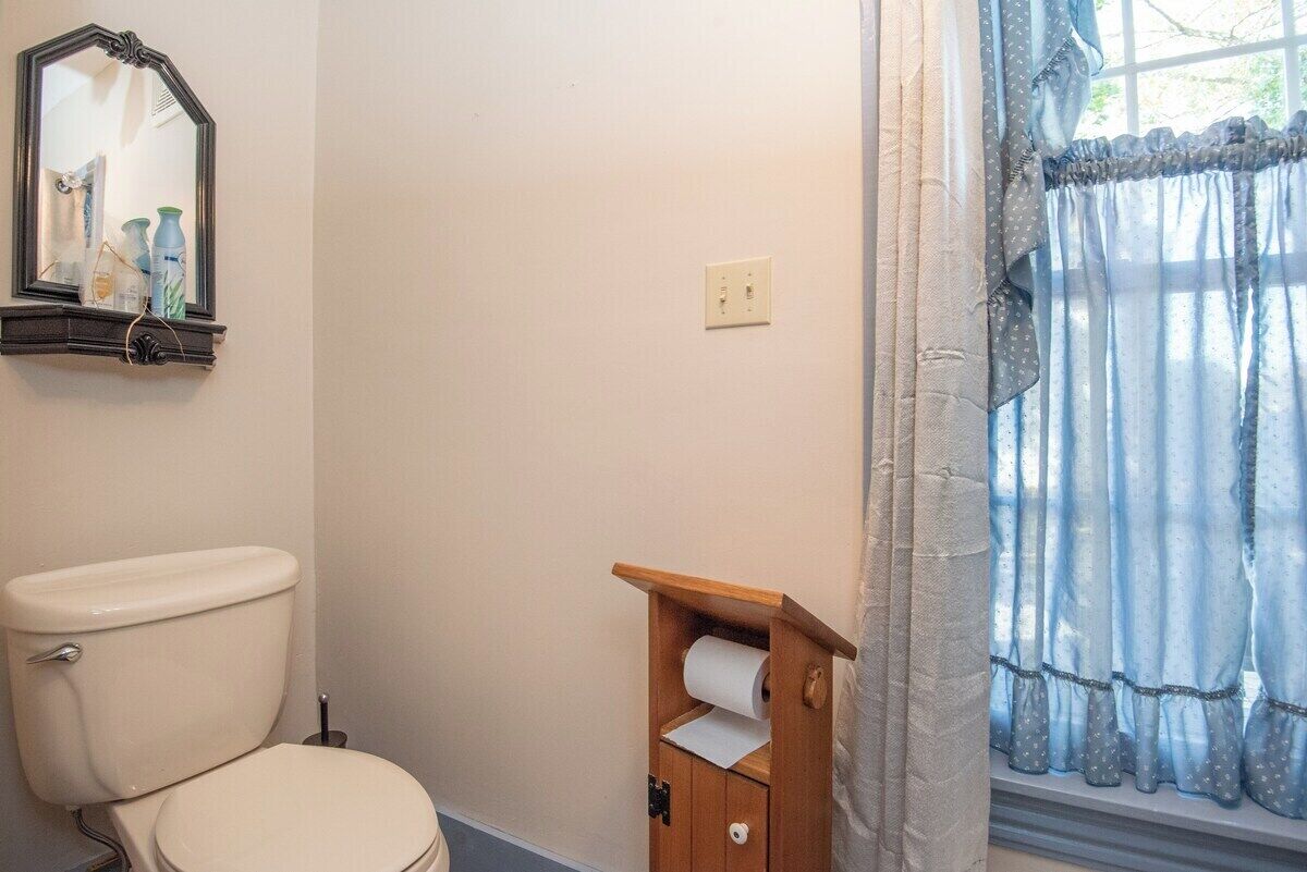 A bathroom with a toilet, a wooden shelf with toilet paper, a mirror, and a window with curtains.