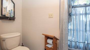 A bathroom with a toilet, a wooden shelf with toilet paper, a mirror, and a window with curtains.