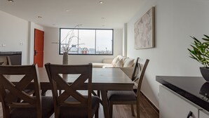 A modern dining area with a wooden table and chairs, a sofa, and a large window offering a city view. - Departamento En La Playa De Estreno Con Piscina (Punta Hermosa)