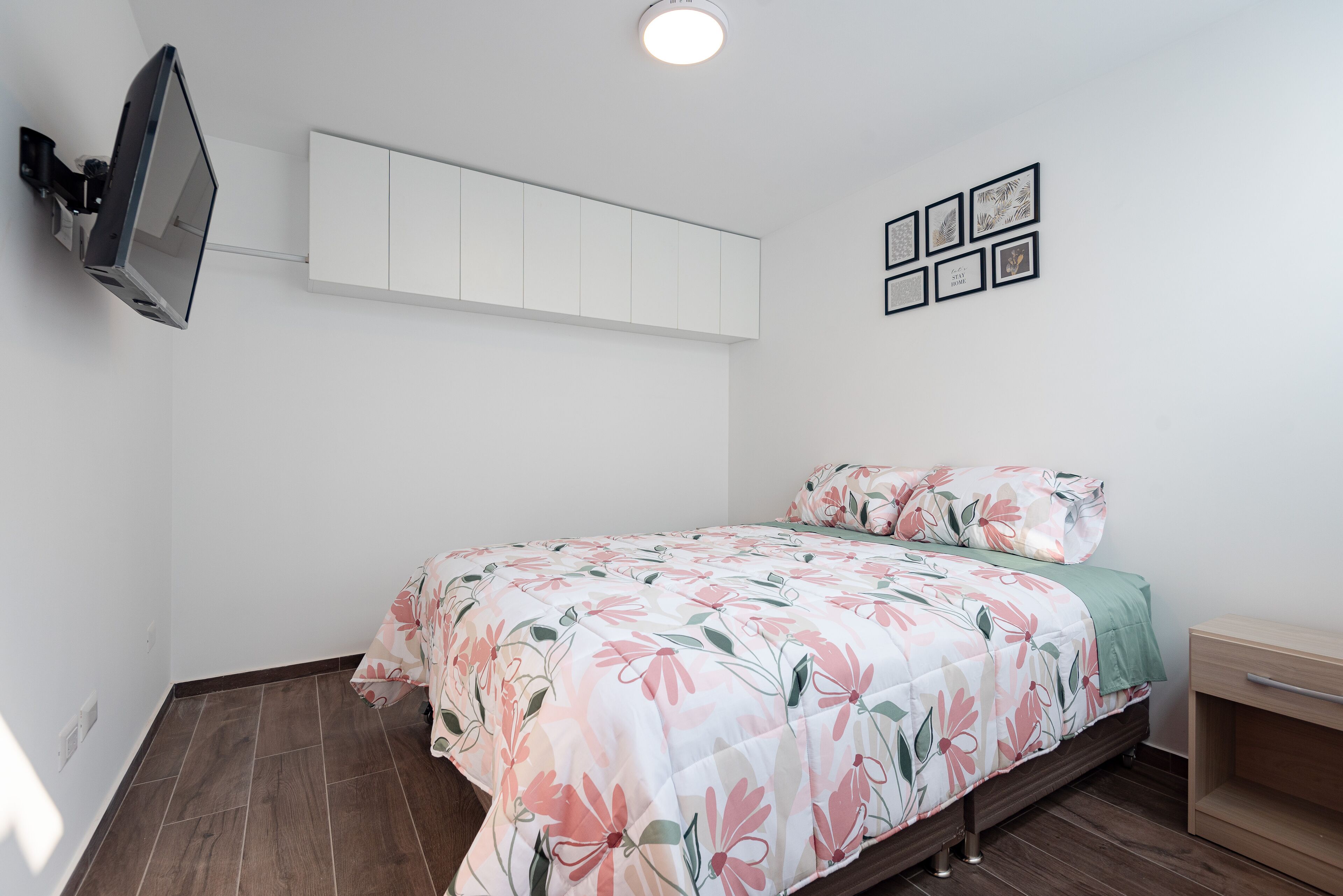 Een slaapkamer met een bed, een aan de muur bevestigde televisie en een klein houten nachtkastje.