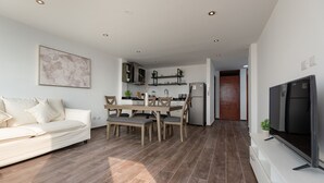 A modern living room with a sofa, dining table, and chairs. There is a kitchen area with a refrigerator, microwave, and cabinets. A TV is mounted on the wall. - Departamento En La Playa De Estreno Con Piscina (Punta Hermosa)