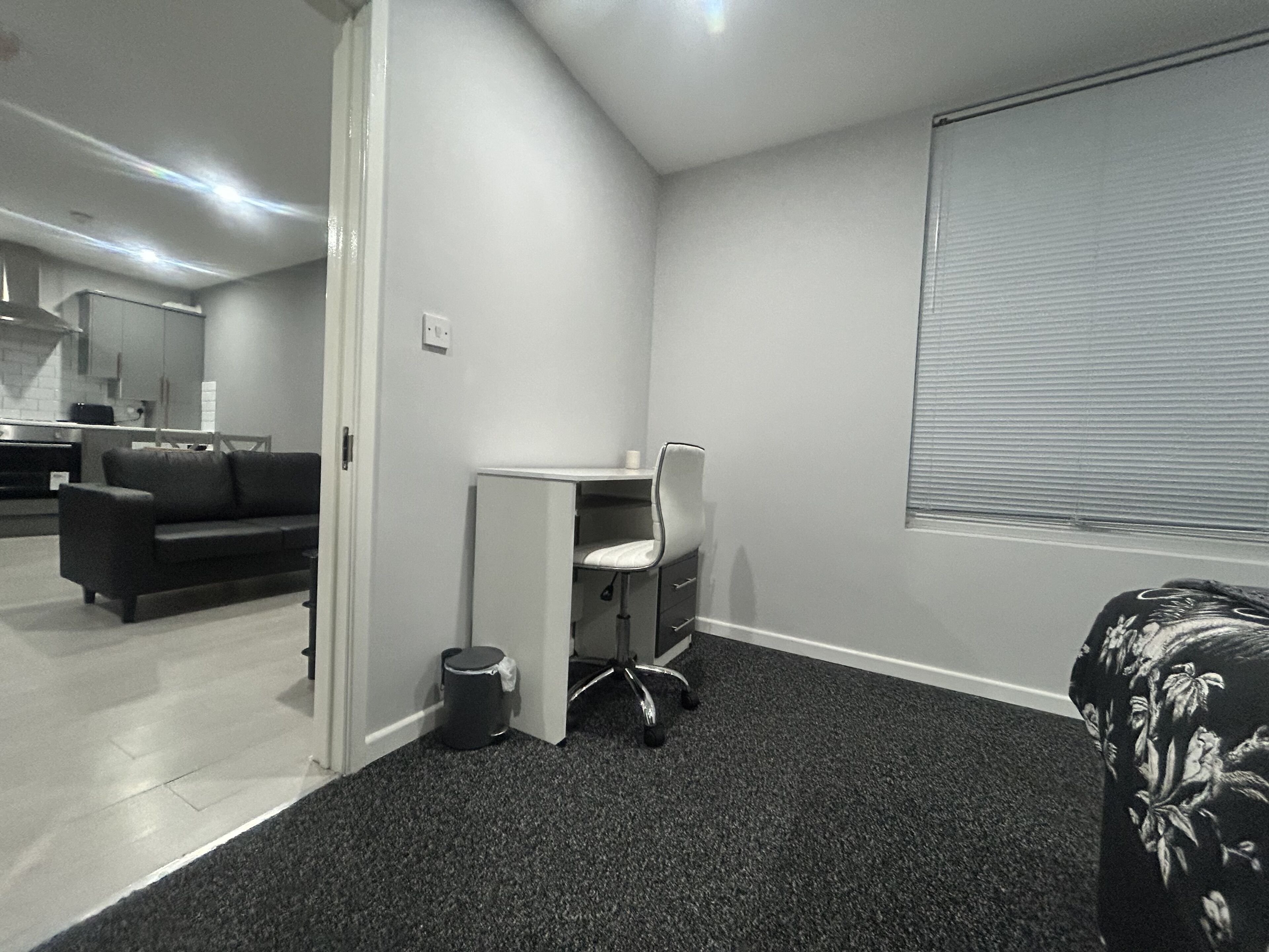 Ein kleines Zimmer mit Schreibtisch, einer kleinen Mülltonne, einer Couch und einem Fenster mit Jalousien.