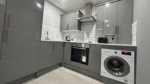 A compact kitchen with grey cabinets, a built-in oven, a washing machine, and a coffee maker. - Private Luxury Apartment - Free Parking & Wi-fi (Coventry)