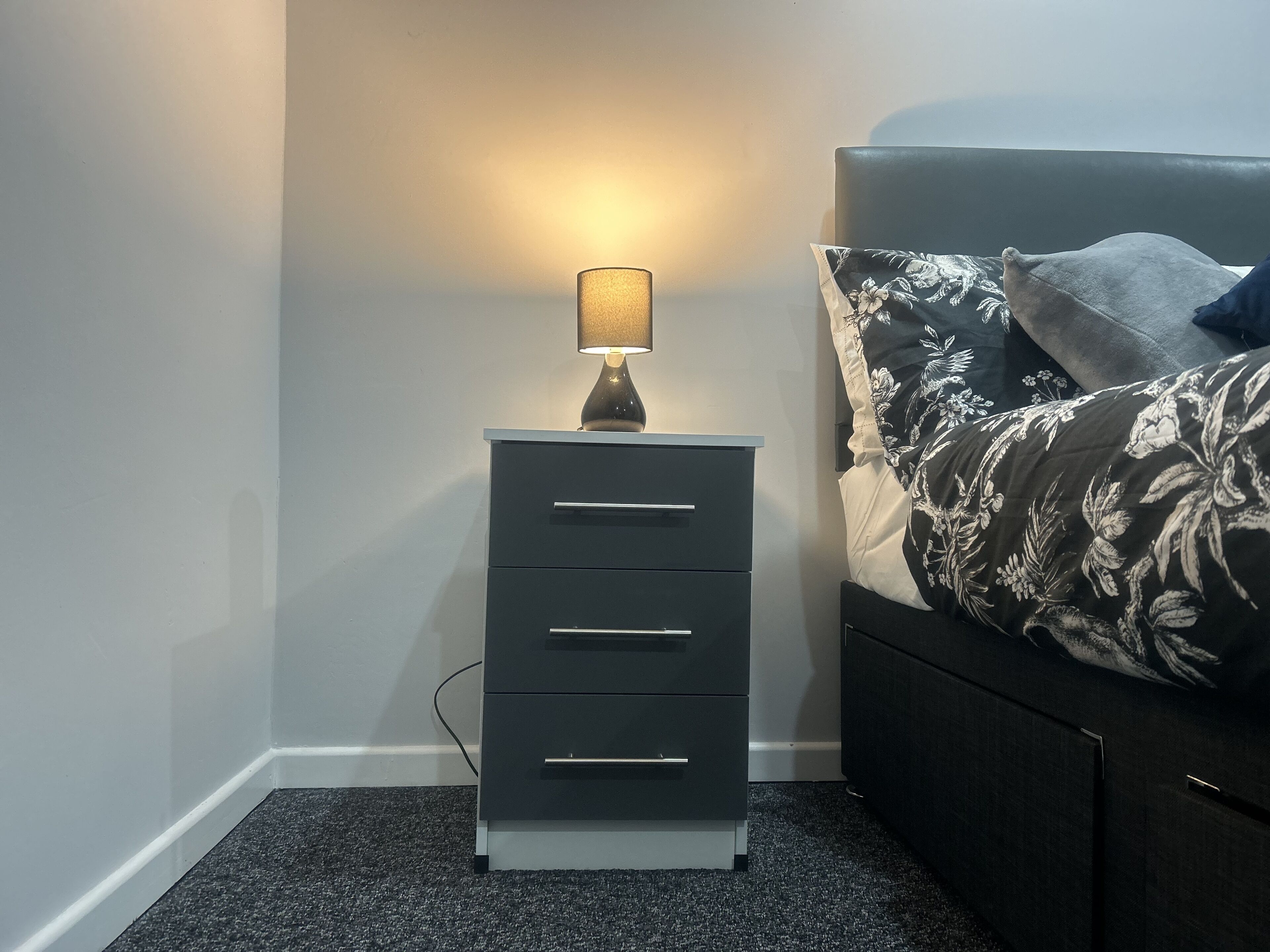 Una mesita de noche pequeña con una lámpara, una cama oscura con ropa de cama estampada con motivos florales y una alfombra gris.