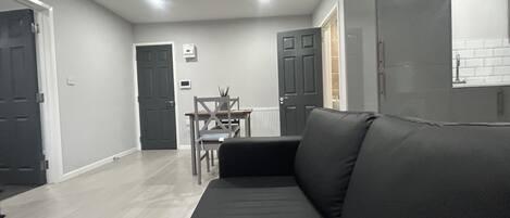 Una sala de estar moderna con un sofá negro, una mesa de comedor con sillas y una zona de cocina con armarios.