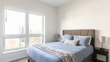 Une chambre avec une grande fenêtre, un lit avec une literie bleue et deux tables de chevet avec des lampes.