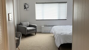 A bedroom with a bed, a gray armchair, a small table, a radiator, and a window with blinds.
