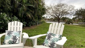 Deux chaises Adirondack blanches, dotées de coussins à motifs, sont placées sur une terrasse en béton, avec une pelouse et des arbres en arrière-plan.