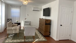 Ein Wohnzimmer mit einem beigen Sofa, einem gemusterten Sessel, einer Holzkommode und einem an der Wand montierten Fernseher.