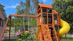 A wooden play structure with a slide and climbing area, surrounded by a garden with pink flowers and a lawn. - Villa Provençale Avec Piscine (Le Cannet-des-Maures)