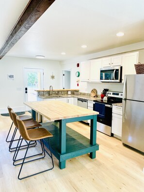 A modern kitchen with a wooden table, bar stools, white cabinets, a stainless steel refrigerator, and a microwave.