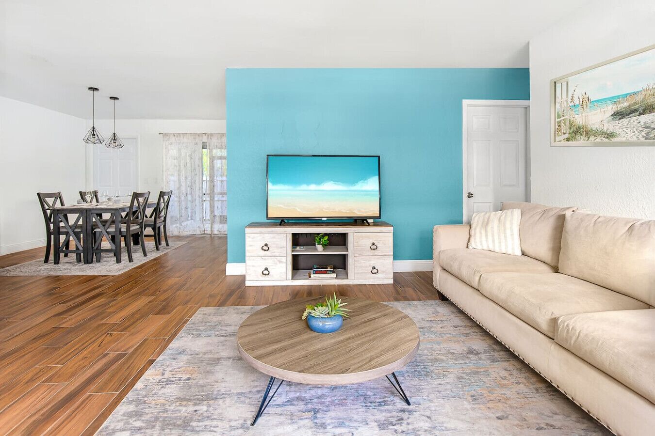 A living room with a teal accent wall, a beige sofa, a wooden coffee table with a plant, a TV on a white cabinet, and a dining area with a wooden table and chairs.