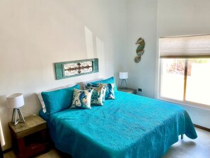 A bedroom with a bed, pillows, a nightstand, a lamp, a wall decoration, and a window.