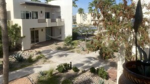 A modern building with a flat roof, large windows, and a balcony. There is a garden area with various plants and a palm tree.