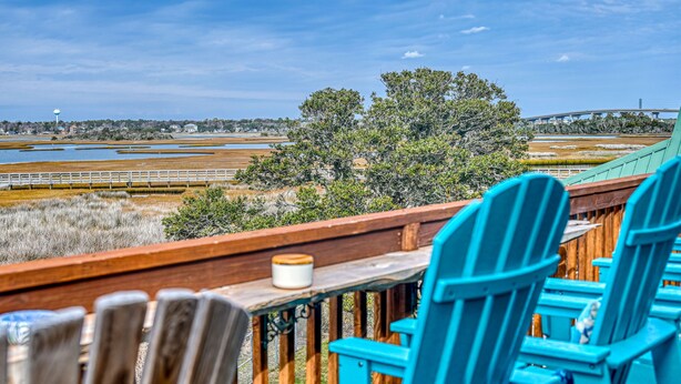 A wooden deck with blue chairs and a table, overlooking a coastal landscape with trees and a bridge. - Wonder the Sea:Sweeping Intracoastal Views, Walk to Beach, Hot Tub, Pets Welcome (Surf City)