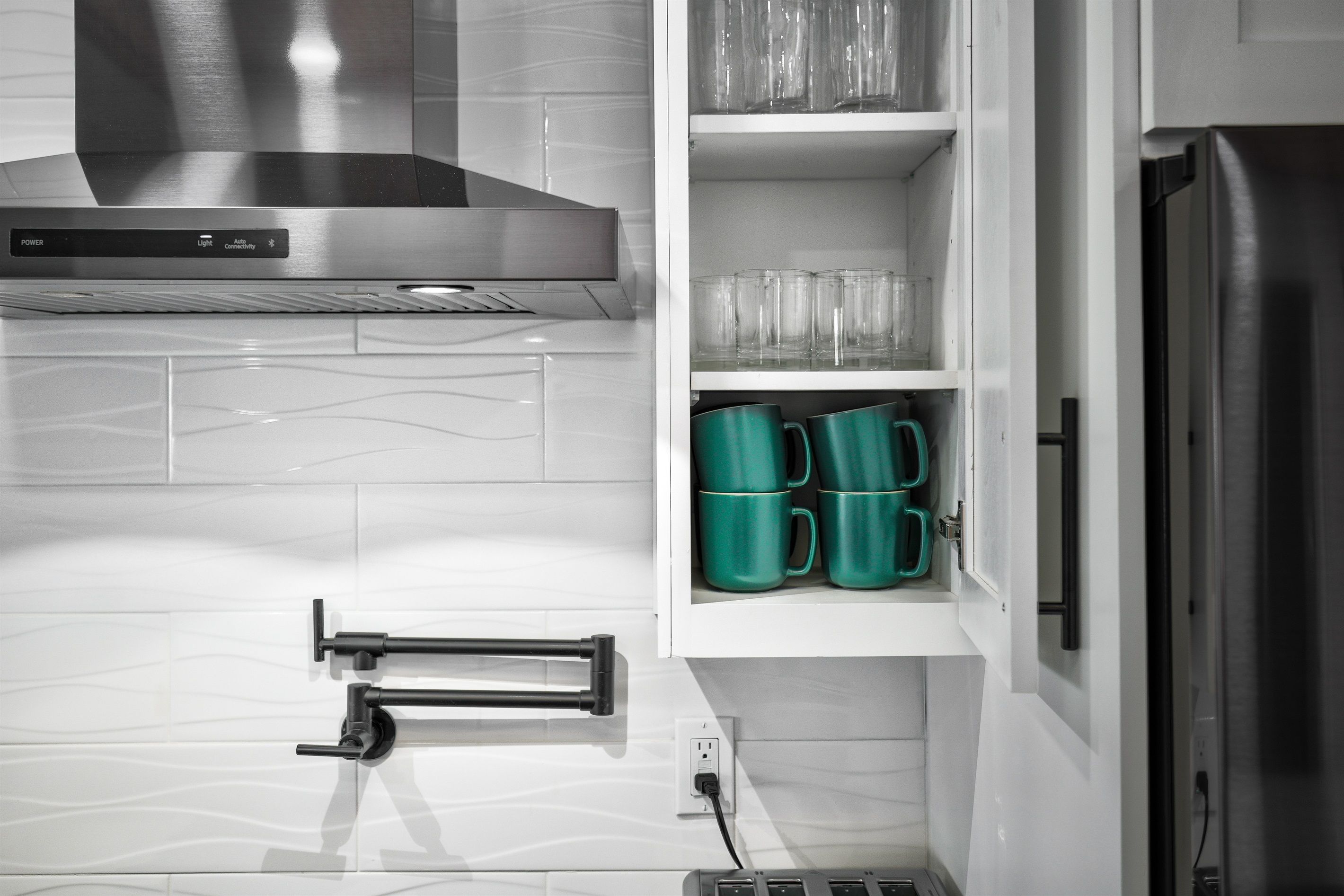 A kitchen with a stainless steel range hood, a built-in oven, and a refrigerator. There are green mugs on a shelf and a power outlet on the wall.