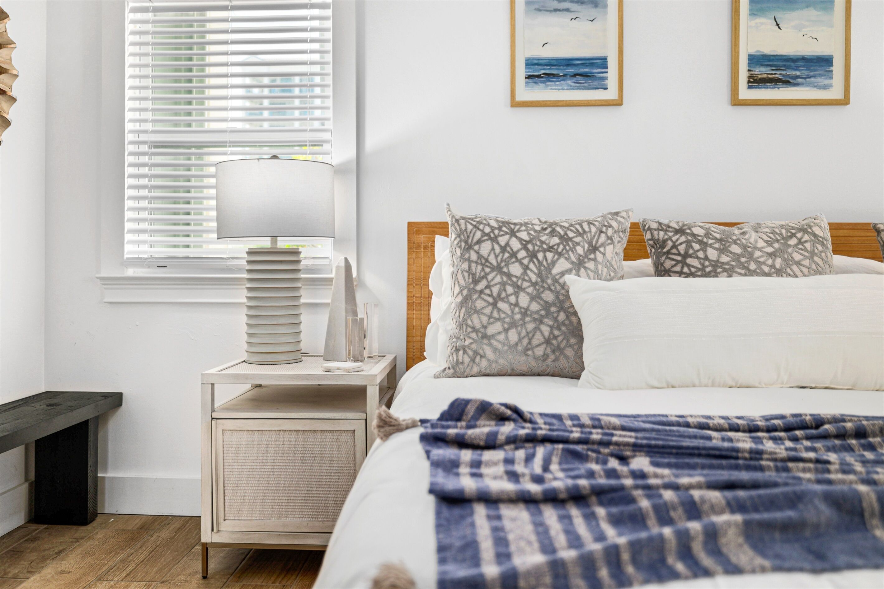 A bedroom with a bed, a bedside table with a lamp, a nightstand, and a window with blinds. There are two framed pictures on the wall.