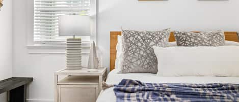 A bedroom with a bed, a bedside table with a lamp, a nightstand, and a window with blinds. There are two framed pictures on the wall.